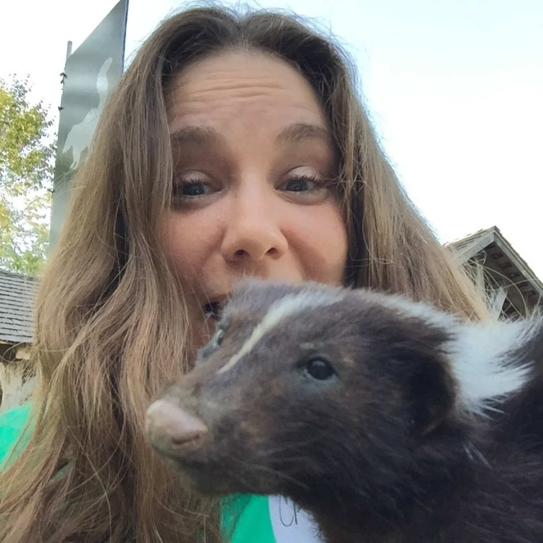 Photo of a white woman looking at the camera. She has long brown hair framing her face and is wearing a green shirt. A taxidermy skunk is being held in front of her face. 
